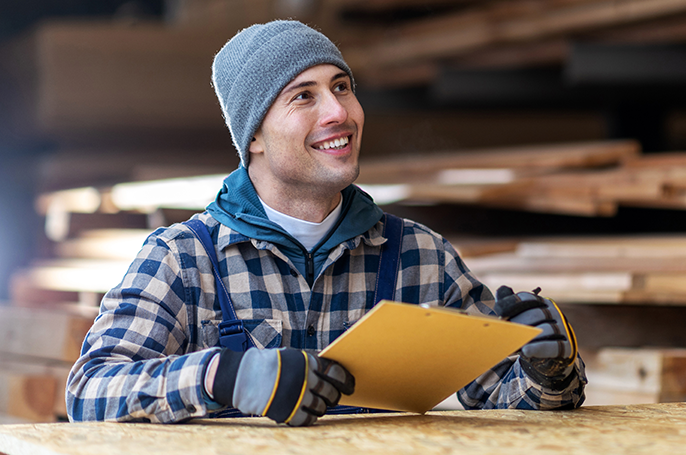 Lächelnder Handwerker in Holzlager mit Klemmbrett in der Hand.